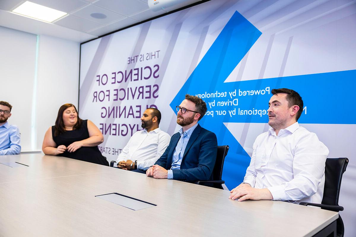 A group of people sitting by a table in a Mitie meeting room with 'This is the Science of Service for Cleaning & Hygiene' on the wall behind them