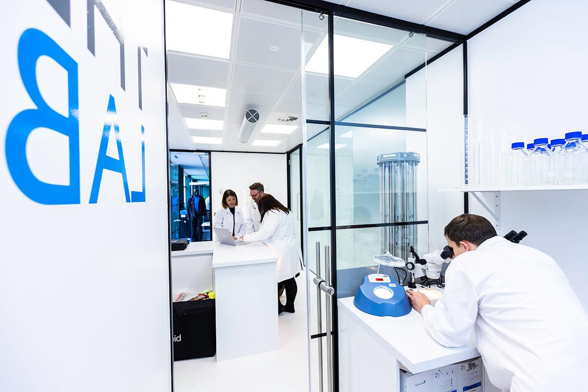 A group of people wearing white lab coats, one looking through a microscope, in a white-walled set of rooms called The Lab in the Mitie Cleaning and Hygiene Centre of Excellence