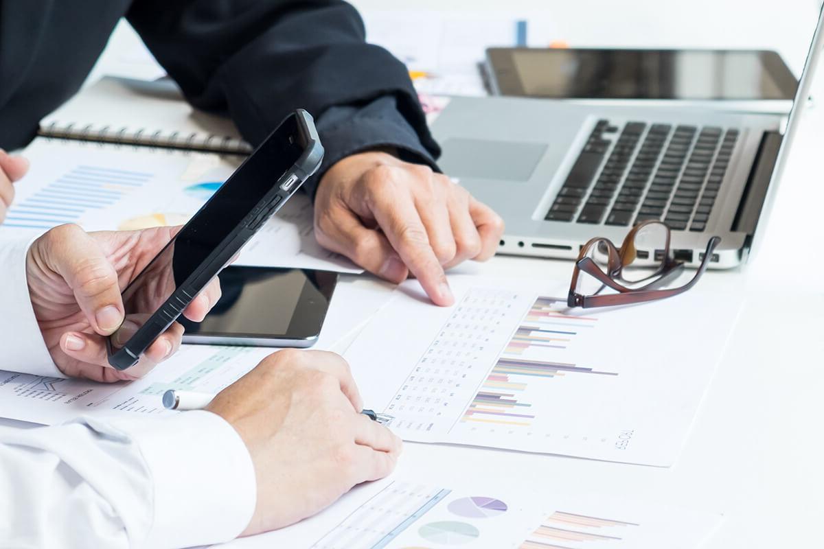 Close up of two people's hands holding smartphones and pointing at charts printed out on paper laying on the desk